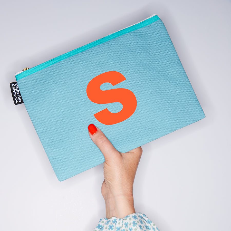 HAND HOLDING LARGE COTTON TURQUOISE PURSE WITH AN ORANGE LETTER S ON IT