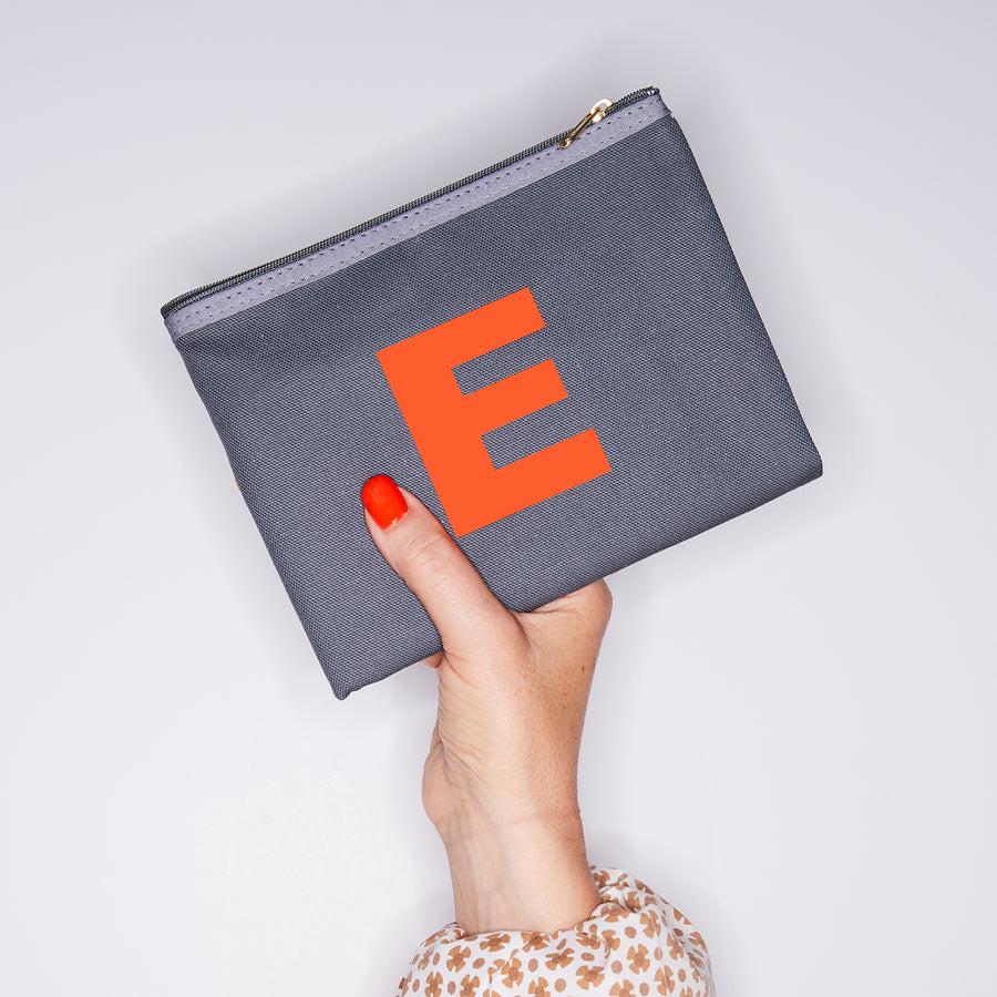 HAND HOLDING MEDIUM COTTON GREY PURSE WITH AN ORANGE LETTER E ON IT