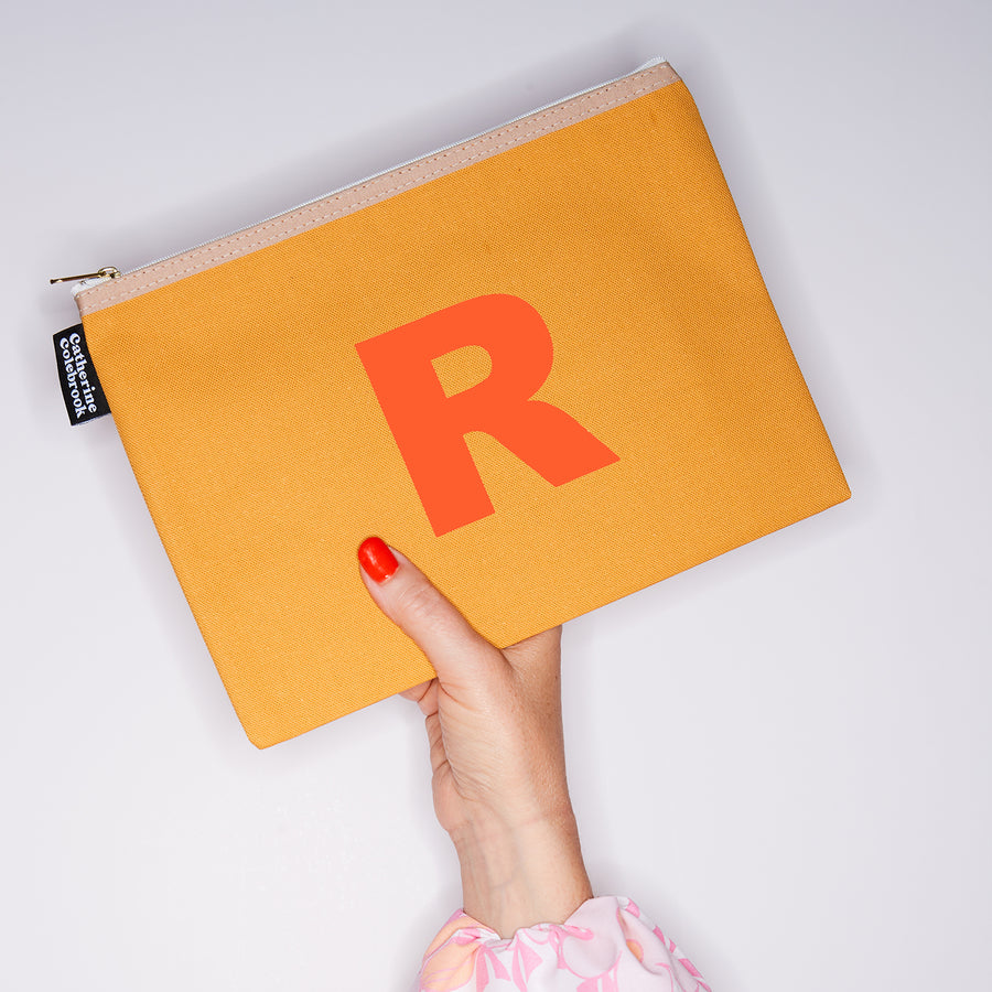 HAND HOLDING LARGE COTTON MUSTARD PURSE WITH AN ORANGE LETTER R ON IT