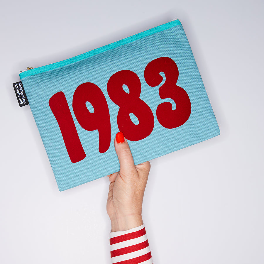 HAND HOLDING TURQUOISE COTTON YEAR PURSE WITH A RED 1983 ON IT