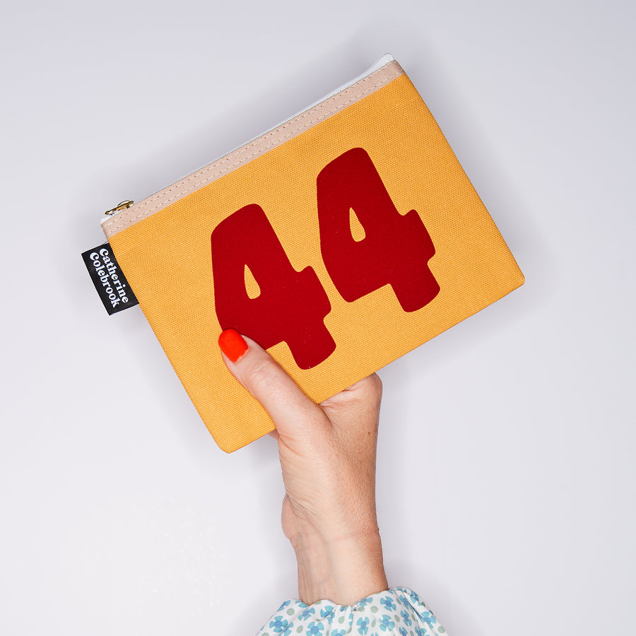 HAND HOLDING MUSTARD COTTON PURSE WITH A RED NO 44 ON IT